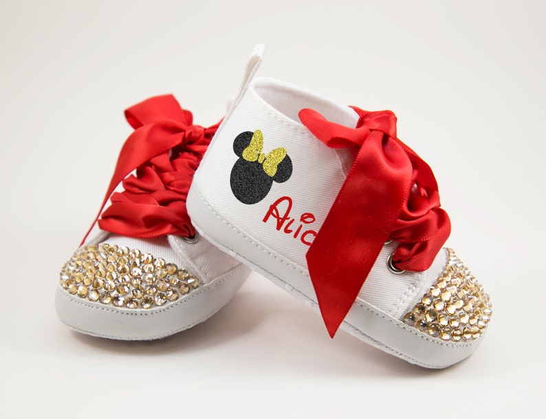 May include: White baby shoes with red satin bows and a black and gold Minnie Mouse design. The shoes are decorated with clear rhinestones and the name "Alice" is written on the side.
