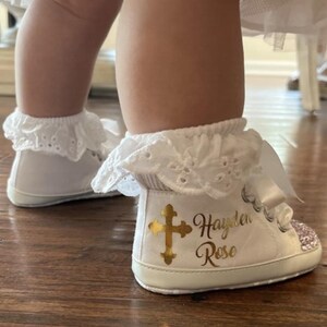 Pode incluir: Sapatos de beb&eacute; brancos com uma cruz dourada e o nome "Hayden Rose" escrito neles. Os sapatos t&ecirc;m uma borda de renda branca e uma fita branca.