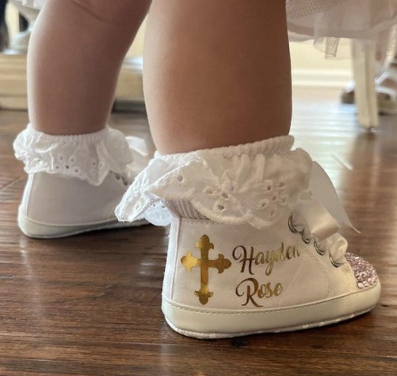 Pode incluir: Sapatos de beb&eacute; brancos com uma cruz dourada e o nome "Hayden Rose" escrito neles. Os sapatos t&ecirc;m uma borda de renda branca e uma fita branca.