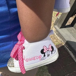 May include: White baby shoes with a Minnie Mouse graphic and the name "Kennedy" in pink. The toe of the shoe is embellished with small, clear rhinestones, and the shoe has a pink polka dot ribbon.