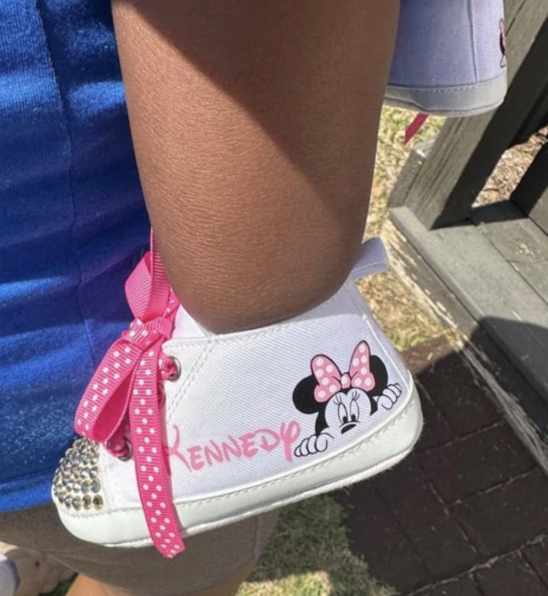 May include: White baby shoes with a Minnie Mouse graphic and the name "Kennedy" in pink. The toe of the shoe is embellished with small, clear rhinestones, and the shoe has a pink polka dot ribbon.