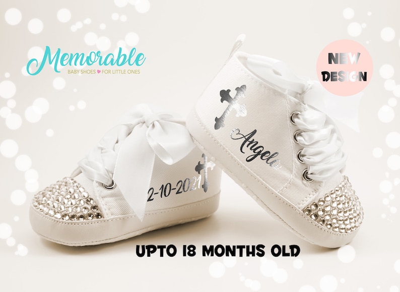 Pode incluir: Sapatos de beb&eacute; brancos com uma cruz prateada e o nome "Angela" bordado. Os sapatos s&atilde;o decorados com strass e t&ecirc;m um la&ccedil;o de cetim branco. Os sapatos s&atilde;o para beb&eacute;s at&eacute; 18 meses.