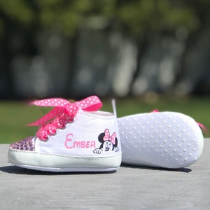 May include: White baby sneakers with pink polka dot laces and a sequined toe. The name "Ember" is printed in pink, along with a Minnie Mouse graphic. The sole is white with small, white, circular grips.