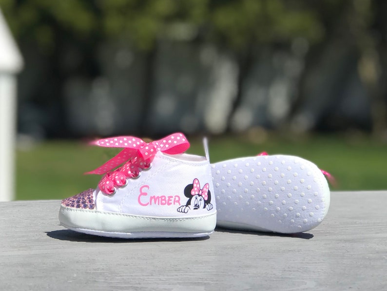 May include: White baby sneakers with pink polka dot laces and a sequined toe. The name "Ember" is printed in pink, along with a Minnie Mouse graphic. The sole is white with small, white, circular grips.
