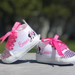 May include: A pair of white baby sneakers with pink polka dot laces and a pink bow. The shoes feature a Minnie Mouse graphic and the name "Ember" in pink. The toe caps are embellished with pink rhinestones.