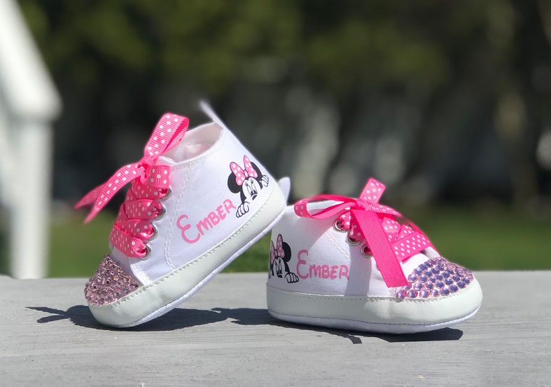 May include: A pair of white baby sneakers with pink polka dot laces and a pink bow. The shoes feature a Minnie Mouse graphic and the name "Ember" in pink. The toe caps are embellished with pink rhinestones.