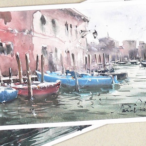 May include: Watercolour paintings depicting a Venetian canal scene with gondolas. The artwork shows buildings in red and white, with blue and red boats in the water. A dried eucalyptus branch is in the foreground.