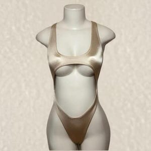 May include: A champagne-colored one-piece swimsuit featuring a high-cut leg design, a scoop neckline, and a large circular cutout detail at the midsection. The garment is crafted from a smooth, satin-like fabric with a subtle sheen throughout.