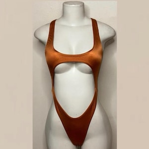 May include: A metallic copper-colored one-piece swimsuit featuring a scoop neckline, thick shoulder straps, and a large circular cutout at the midsection that exposes the torso, finished with a high-cut leg design and a smooth, shiny fabric.