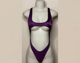 May include: A deep purple one-piece swimsuit featuring a high-cut leg design, a scoop neckline, and a large circular cutout at the midsection. The garment has thick shoulder straps and a racerback style, crafted from a smooth, solid-colored fabric.