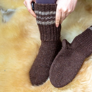 Puede incluir: Un par de calcetines de punto marrón con un detalle de rayas beige. Los calcetines se muestran sobre una superficie peluda.