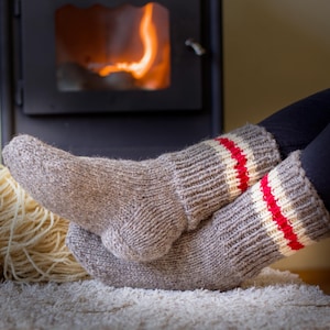 May include: A pair of gray wool socks with red and white stripes. The socks are worn by a person sitting in front of a fireplace.