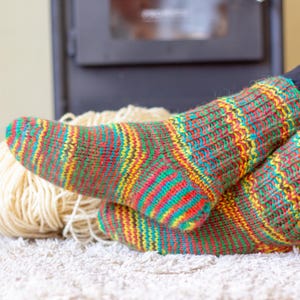Puede incluir: Primer plano de un par de calcetines de punto coloridos con una vibrante mezcla de rayas rojas, amarillas, verdes y azules. Los calcetines descansan sobre una superficie blanca y texturizada junto a una madeja de lana color crema. Los calcetines parecen hechos a mano.