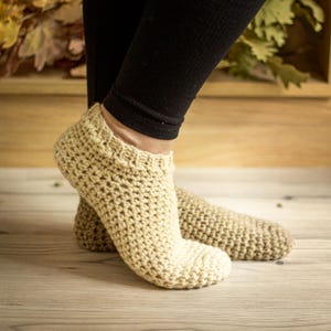 May include: A pair of beige crochet socks. The socks are thick and textured, with a ribbed cuff and a rounded toe. The socks are worn on a person's feet, with black leggings visible above the socks. The background is a wooden surface.