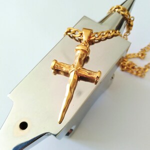 May include: A gold-toned cross pendant necklace with a textured, pointed design. The necklace is on a silver-toned chain and is displayed on a metal surface.