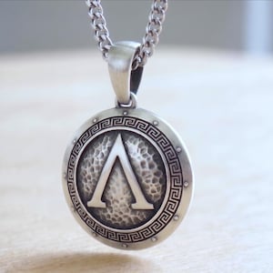 May include: Silver-toned pendant necklace featuring a round shield design with a hammered texture. The pendant showcases a prominent silver letter "Λ" within a circular border with a Greek key pattern. The necklace has a curb chain.
