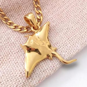 Può includere: Un ciondolo color oro a forma di manta, caratterizzato da una superficie liscia e riflettente e da una coda lunga e sottile. Il ciondolo è fissato a una catena a maglie gourmette abbinata, con una finitura lucida che cattura la luce.