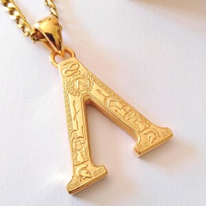 May include: A gold-toned pendant shaped like the Greek letter Lambda, suspended from a matching chain. The surface of the letter is engraved with detailed motifs including a Spartan helmet, a spear, a shield, and a coiled snake design.
