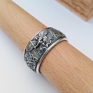 May include: A silver ring with detailed relief carvings. The ring features a central winged figure with a sword, surrounded by intricate knotwork and other symbolic elements. The ring is displayed on a wooden dowel.