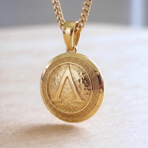Könnte beinhalten: Goldfarbene Halskette mit Anhänger. Der runde, strukturierte Anhänger zeigt den griechischen Buchstaben Lambda. Er ist von einem dekorativen Rand umgeben und hängt an einer passenden Goldkette.