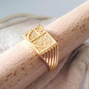 May include: A gold-colored ring with a rectangular face featuring a textured design and a curved symbol. The ring is set on a wooden dowel, showcasing its intricate details and polished finish. The ring's band has vertical grooves.