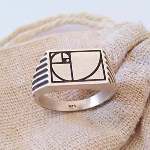 Könnte beinhalten: Ein silberner Ring mit rechteckiger Oberfläche, der ein schwarz-weißes geometrisches Design aufweist. Der Ring hat eine glatte, polierte Oberfläche und die Zahl "925" ist auf dem Band sichtbar. Der Ring ist vor einem neutralen Hintergrund platziert.