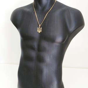 May include: A gold-colored pendant necklace featuring the Ukrainian Tryzub emblem. The pendant hangs from a delicate chain and is displayed on a black mannequin torso. The necklace is a symbol of Ukrainian heritage.