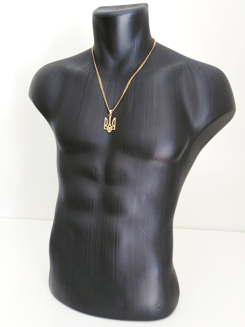 May include: A gold-colored pendant necklace featuring the Ukrainian Tryzub emblem. The pendant hangs from a delicate chain and is displayed on a black mannequin torso. The necklace is a symbol of Ukrainian heritage.