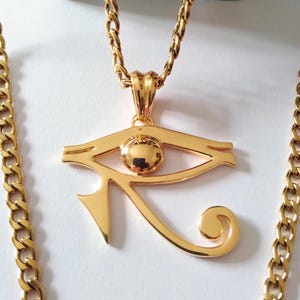 May include: A gold chain necklace with a large gold pendant featuring the Eye of Horus symbol. The pendant is detailed with a raised eye and a curved wing.