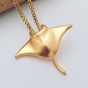 Könnte beinhalten: Eine goldfarbene Anhängerkette mit einem Mantarochen-Design. Der Anhänger hat eine strukturierte Oberfläche und hängt an einer filigranen Kette. Der Mantarochen-Anhänger ist etwa 5 cm breit.