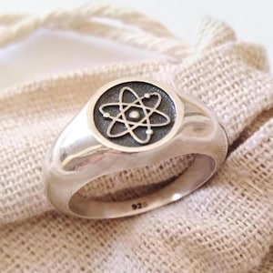 May include: A silver ring with a polished finish. The ring features a circular emblem with a detailed atom design in the center. The ring is set against a neutral fabric background, highlighting the ring's metallic sheen.