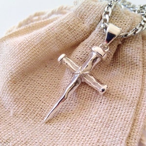 May include: Silver nail-shaped cross pendant on a silver chain. The pendant is on a beige fabric background.