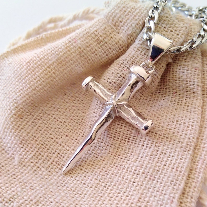 May include: Silver nail-shaped cross pendant on a silver chain. The pendant is on a beige fabric background.