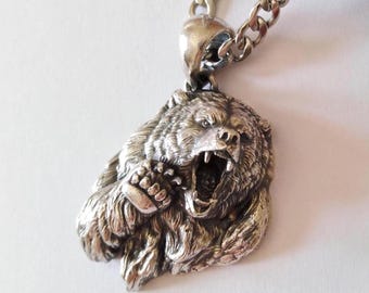 May include: A silver-toned metal pendant shaped like the head and front paw of a roaring bear with detailed fur texture and an open mouth showing teeth. The pendant is attached to a thick silver-toned metal chain link necklace.