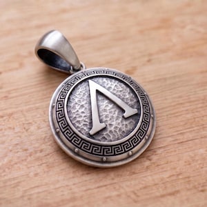 May include: Silver-toned pendant featuring a circular design with a textured surface and a Greek Lambda symbol in the center. The pendant has a decorative border with a key pattern and a loop for a chain. The pendant is displayed on a wooden surface.