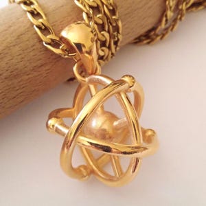 Pode incluir: Um colar com pingente dourado com um design esférico dentro de uma série de aros que se cruzam. O pingente pende de uma grossa corrente dourada. O colar é exibido em um fundo claro.