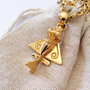 Puede incluir: Un collar con colgante dorado con un diseño de rana estilizado. El colgante tiene una forma tridimensional detallada con formas geométricas y acentos en espiral. El collar se muestra sobre una bolsa de tela beige, con una cadena dorada.