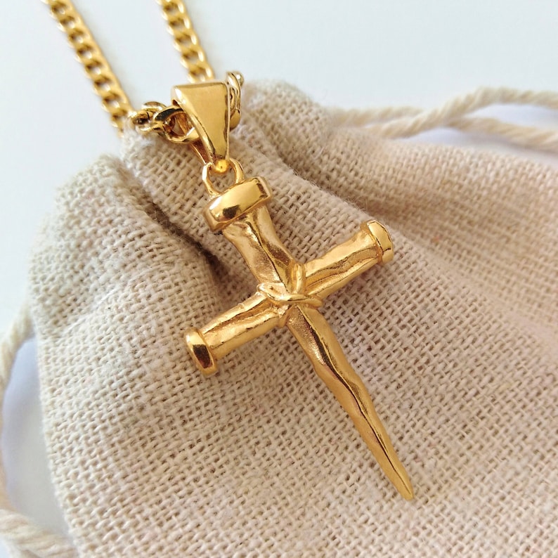 May include: Gold chain necklace with a gold cross pendant. The cross is shaped like a nail with a textured surface.
