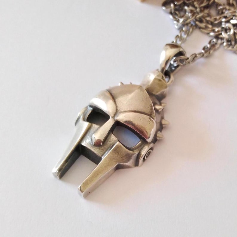 May include: Silver pendant necklace featuring a detailed, three-dimensional gladiator helmet with spikes. The helmet is silver and has a textured surface.