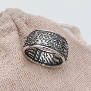 May include: A silver ring with an intricate, engraved pattern around the band. The ring features a detailed design with a repeating pattern and text inside the band. The ring is displayed on a beige fabric surface.