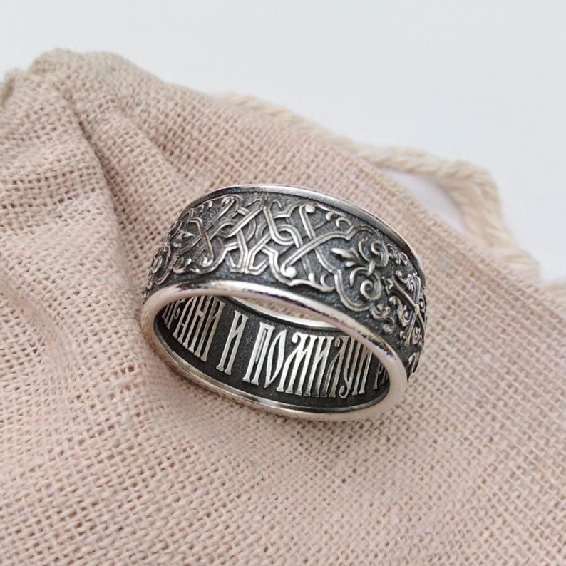 May include: A silver ring with an intricate, engraved pattern around the band. The ring features a detailed design with a repeating pattern and text inside the band. The ring is displayed on a beige fabric surface.