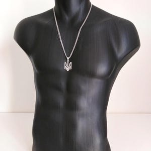 May include: A silver-colored necklace featuring a pendant in the shape of the Ukrainian coat of arms, the Tryzub. The pendant hangs from a chain necklace. The necklace is displayed on a black mannequin.