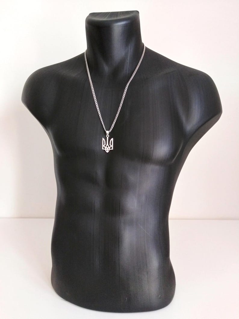 May include: A silver-colored necklace featuring a pendant in the shape of the Ukrainian coat of arms, the Tryzub. The pendant hangs from a chain necklace. The necklace is displayed on a black mannequin.