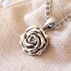 May include: A silver-colored rose pendant necklace. The pendant is a detailed, three-dimensional rose with intricate petal detailing. The necklace chain is a thick, curb style. The jewelry is displayed on a textured, light-colored fabric.