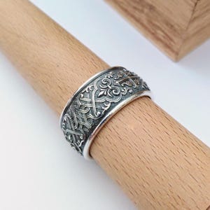 May include: A silver-colored ring with an intricate, detailed pattern of interwoven lines and floral designs. The ring is wide and sits on a wooden dowel, showcasing its ornate craftsmanship. The ring's design features a vintage or antique aesthetic.