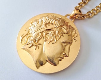 Op de afbeelding: Een gouden ketting met een ronde hanger met een gedetailleerd profiel van een mannengezicht. De hanger is in reliëf, waardoor hij een 3D-effect heeft.
