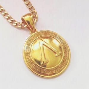 Könnte beinhalten: Eine goldfarbene Anhängerkette mit einem runden Medaillon, das in der Mitte ein erhabenes griechisches Lambda-Symbol zeigt. Das Medaillon ist von einem dekorativen Mäander-Muster umrandet und hängt an einer passenden goldfarbenen Panzerkette.