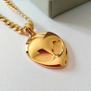 Könnte beinhalten: Eine goldene Kettenkette mit einem goldenen Alienkopf-Anhänger. Der Anhänger ist mit einem Gesicht und Augen verziert.