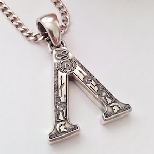 May include: Silver-toned pendant in the shape of the Greek letter lambda, featuring engraved Spartan imagery. The pendant is suspended from a silver chain. The design includes a snake, laurel wreath, spears, shields, and Spartan soldiers.