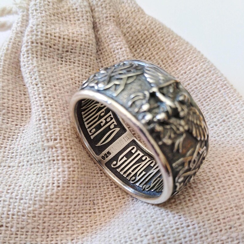May include: A detailed silver ring with engraved religious iconography. The ring features intricate designs, including an eagle and Cyrillic text. The band is marked with "925". The ring is displayed on a beige fabric.
